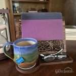 Classic front design wood book stand shown in use with book, tea mug seated on coaster and eyeglasses stored on front coaster.